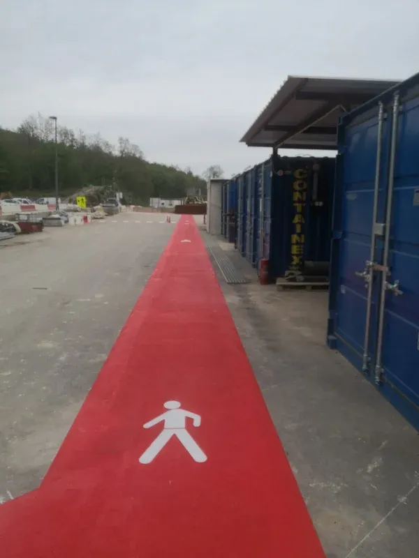 Dugačka crvena pješačka staza s bijelim piktogramom pješaka uz plave kontejnere