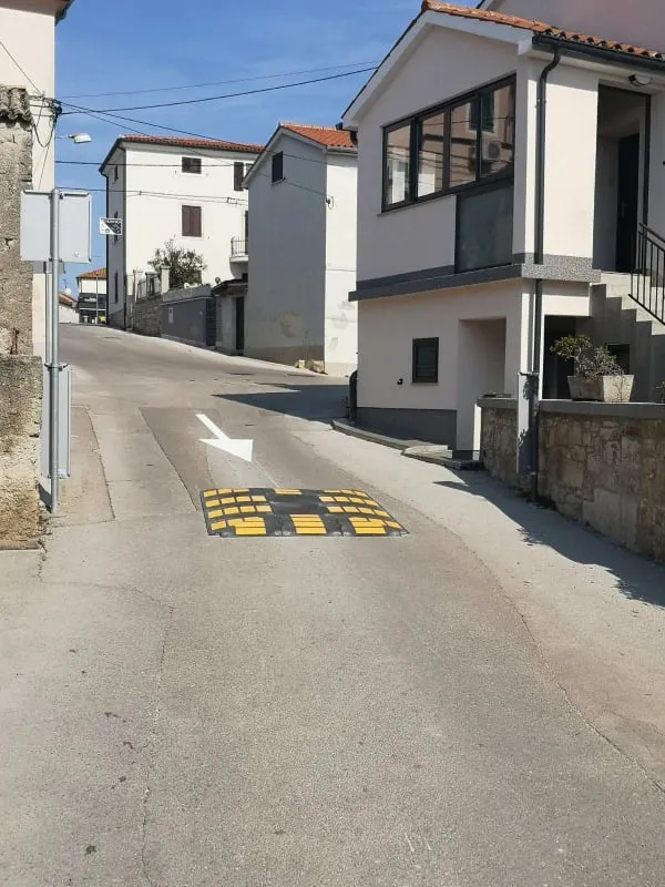Žuto-crni ležeći policajac postavljen na cesti u naselju Pomer uz bijelu strelicu za smjer vožnje.