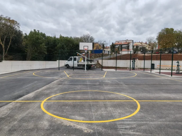 Široki kadar sportskog terena u Puli sa zelenom zaštitnom ogradom i parkiranim radnim vozilom u pozadini.