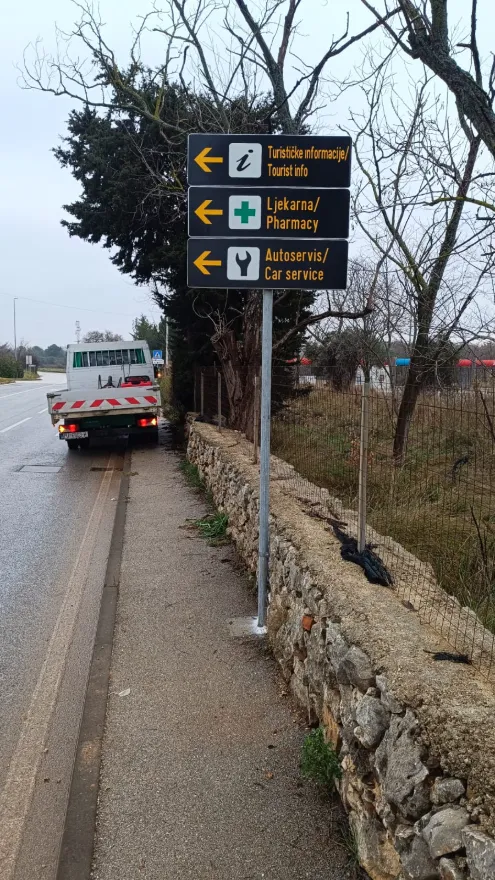 Postavljeni crni putokazi za turističke informacije, ljekarnu i autoservis u Banjolama na pocinčanom stupu.